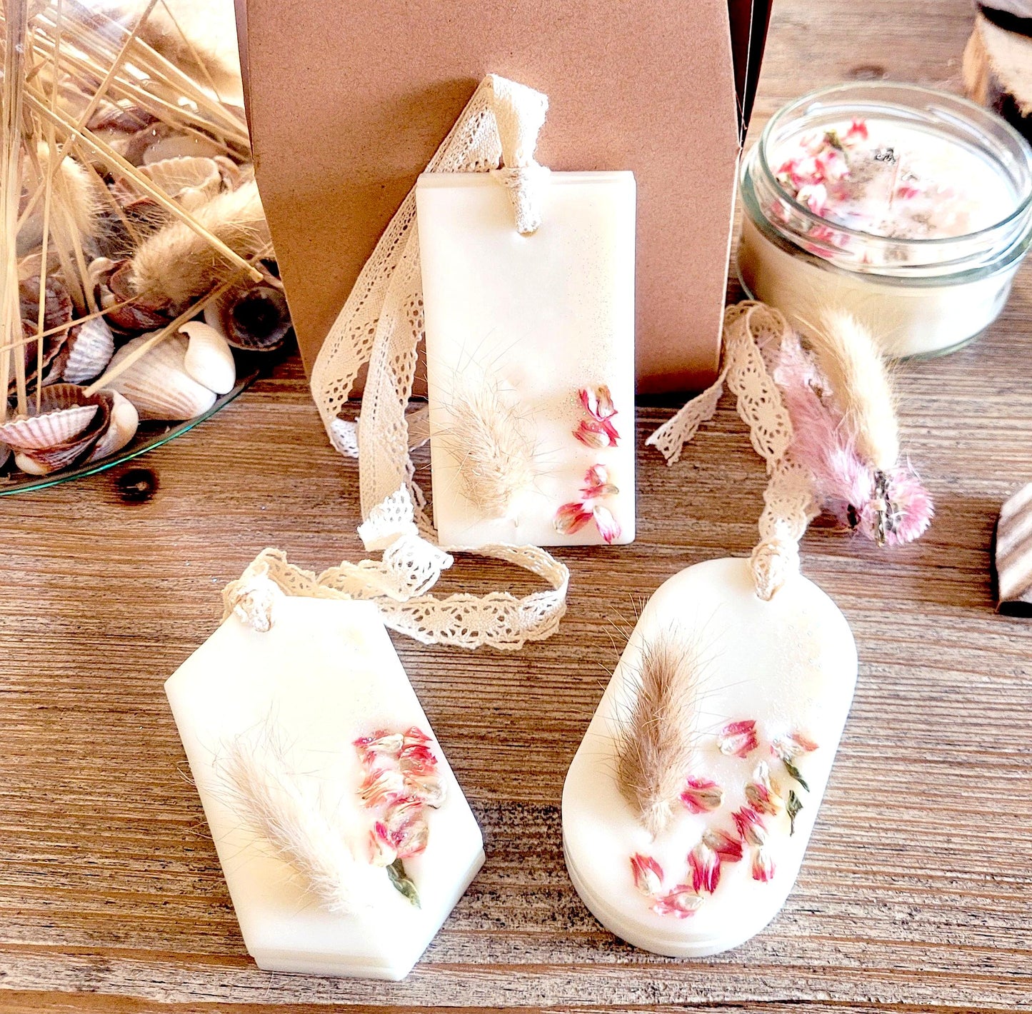 Suspensions parfumées artisanales à la cire végétale, décorées de fleurs séchées roses et de graminées, présentées sur fond en bois naturel.