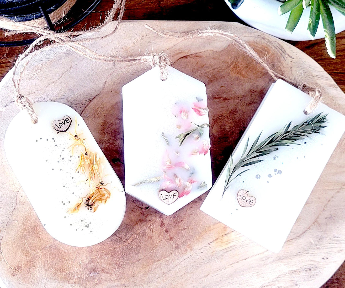 Lot de trois suspensions parfumées artisanales aux formes variées, ornées de fleurs séchées et ficelle naturelle, sur plateau en bois clair.